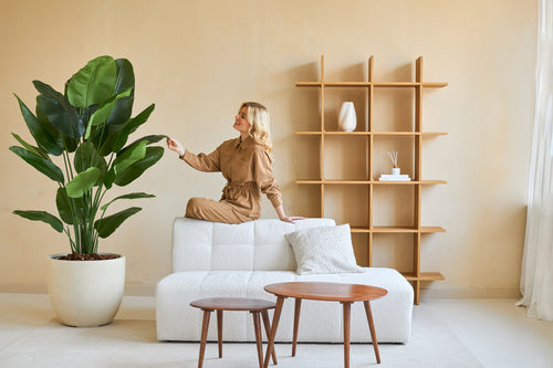 A woman in a brown jumpsuit sits on the back of a white sofa and touches the leaf of an Artificial Strelitzia Real Touch Deluxe 180 cm plant in a minimalist living room with wooden tables and shelves.