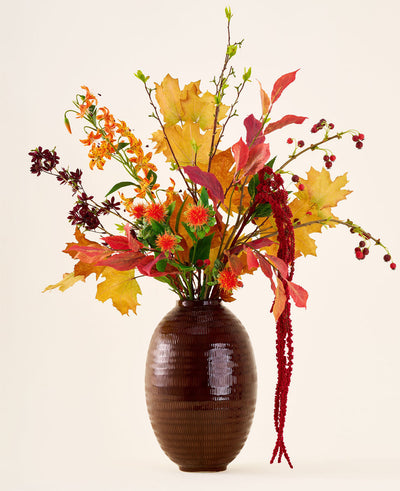 In a brown vase is the 'Rustic Glow' bouquet of silk artificial flowers, with yellow, orange and red leaves, red berries, orange flowers and long, hanging red amaranthus against a neutral background.