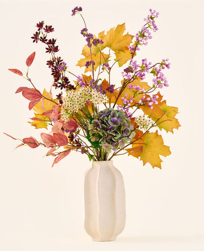 The 'Wild Amethyst' bouquet of silk artificial flowers, with autumn leaves, purple and white flowers and green hydrangeas, is in a white ceramic vase against a plain, light background.