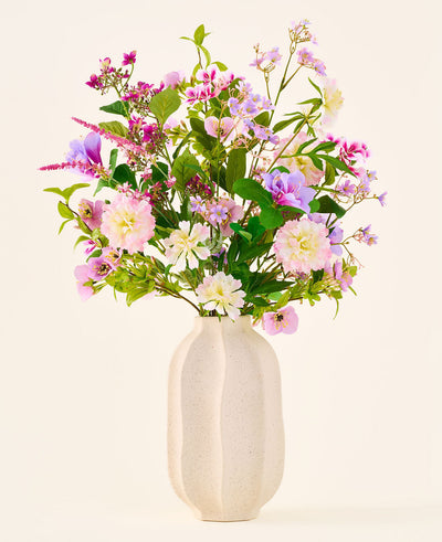 A white ceramic vase holds a colorful bouquet of pink, purple and white flowers from the 'Silk artificial flower bouquet Royal Bloom' collection, accented with green leaves and presented against a plain, light background.