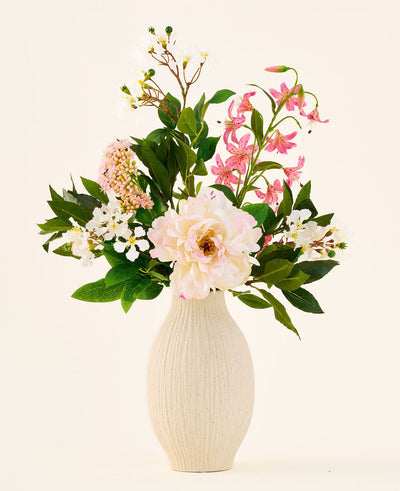In a cream-colored ceramic vase is the 'Light & Lovely' bouquet with artificial silk flowers from Easyplants, consisting of a mixed arrangement of white, pink and light purple silk flowers with green leaves, against a plain, light background.