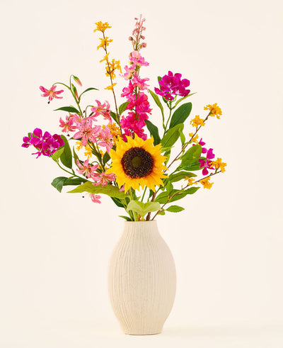 The silk artificial flower bouquet 'Tiny Sunshine' is placed in a beige ceramic vase, consisting of an artificial bouquet of lifelike flowers, such as a large sunflower, pink and magenta blossoms and yellow flowers with green leaves, against a plain light background. This arrangement from Easyplants consists of beautiful silk flowers that add long-lasting beauty to any room.