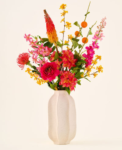 In a white ceramic vase is the 'Summer Blush' bouquet of silk artificial flowers, consisting of pink, yellow and orange flowers with green leaves, against a plain light background.