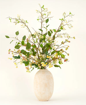 A tall vase made of beige ceramic contains the Silk artificial flower bouquet Wild Apple Blossom XL, with green leafy branches, small yellow flowers and pink berries against a light background.