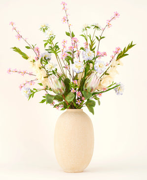 In a vase with a beige structure is the Silk artificial flower bouquet Lots of Love XL, a hand-tied bouquet of pink, white and blue silk flowers with green leaves and long stems against a light background.