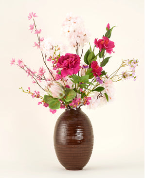 The Pink Reverie silk artificial flower bouquet is in a vase with a brown structure, with pink, magenta and white artificial flowers with green leaves against a plain light background.