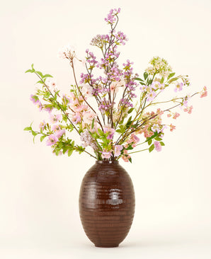 A brown ceramic vase shows the Silk artificial flower bouquet Lilac, a tall and elegant silk bouquet with pink, purple and lilac flowers with green leaves and thin branches against a plain light background.