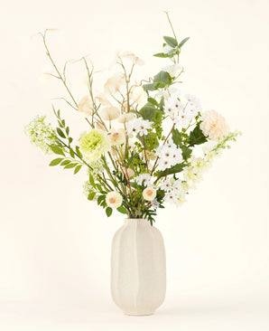 The Claire de Lune silk artificial flower bouquet features delicate white, pale yellow and green flowers in a white ceramic vase, accented by lush greenery against a light background.