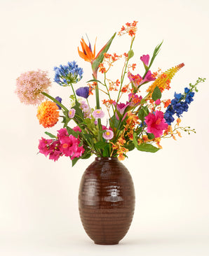 In a vase with a brown structure is the Silk artificial flower bouquet Tropical Firework - an arrangement of orange, pink, purple, yellow and blue silk flowers with green leaves against a plain light background.