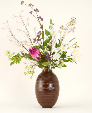 A Serene Bloomfield silk artificial flower bouquet, featuring a large pink protea, white roses, purple and white blossoms and green foliage, stands in a brown ribbed vase against a plain light background.