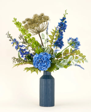 The Mystic Blue Garden silk artificial flower bouquet consists of blue hydrangeas, larkspur, green leaves and dried beige seed heads in a blue ceramic vase - ideal to brighten up your space against a light background.
