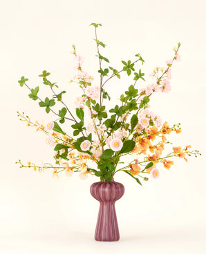 A Blossom Harmony silk artificial flower bouquet is arranged in a mauve vase with pink and peach flowers and green leaves, against a simple light background for an elegant display.