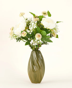 A green, ribbed ceramic vase contains the White Love silk artificial flower bouquet - an arrangement of white and cream-colored flowers with green leaves against a plain light background.