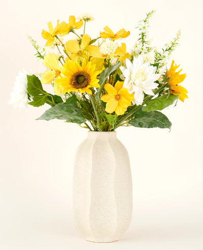 The Sunny Whisper silk artificial flower bouquet, with sunflowers, white dahlias and green leaves in a beige ceramic vase, stands out beautifully against a light background.