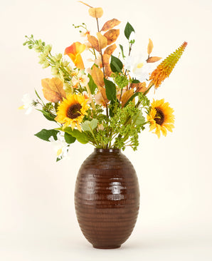 A brown textured vase shows the Amber Light silk artificial flower bouquet, with sunflowers, white and orange flowers and green foliage against a plain background.
