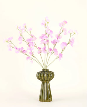 A green glass vase shows the Silk artificial flower bouquet Pea pink with soft green foliage, with several long stems and against a plain off-white background.