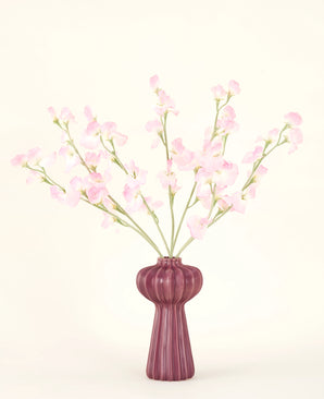 The Silk artificial flower bouquet Pea light pink, with several light pink pea stems in a ribbed burgundy vase, stands against a pale background - ideal for any artificial flower interior.