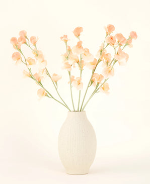 The cream-coloured ceramic vase contains the Silk artificial flower bouquet Pea Peach, with delicate peach and light pink flowers with long green stems against a light, neutral background.