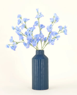A blue ribbed ceramic vase displays the Pea Blue silk artificial flower bouquet, with its delicate light blue artificial flowers against a plain, light background.