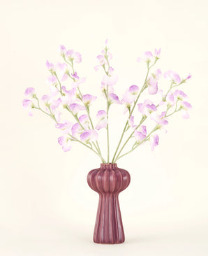 The Silk artificial flower bouquet Pea lilac features long stems of delicate, light purple flowers and green leaves in a mauve ceramic vase with vertical ridges, against a plain, off-white background.