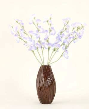A brown, ribbed vase holds the Artificial Silk Flower Bouquet Pea Lavender, with delicate petals and green stems against a plain off-white background for a subtle cottage atmosphere.