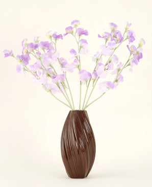 A textured brown vase holds the Artificial Silk Flower Bouquet Pea purple-light purple silk flowers with greenish stems against a plain, light background.
