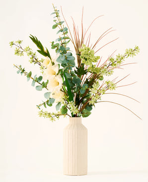 In a beige vase is the bouquet "Wild Ivory" of silk artificial flowers, with green leaves, eucalyptus, white flowers, small green blossoms and long, thin brown grasses against a light background.