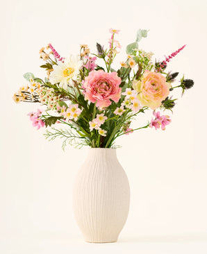 In a white ceramic vase is the 'Backyard Blossom' bouquet of artificial silk flowers, consisting of a beautiful arrangement of pink roses, white daisies and small yellow flowers, made from silk flowers, supplemented with lush green leaves against a plain light background.