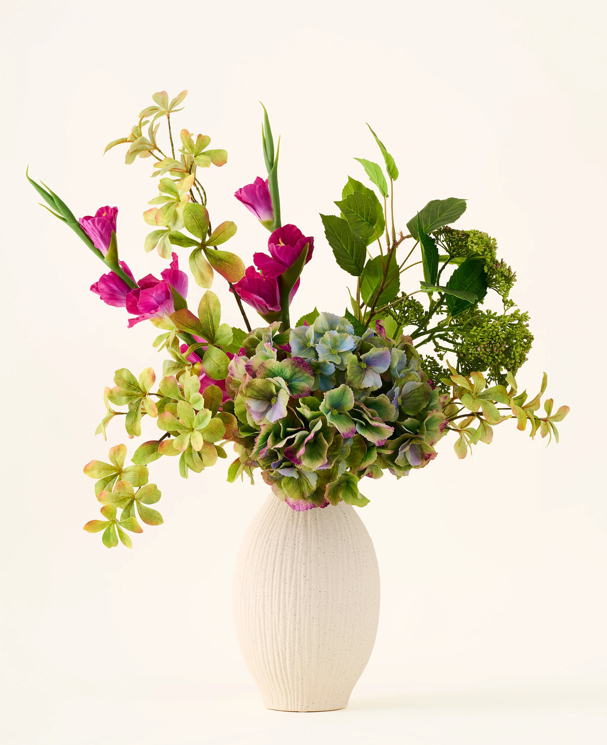 A white textured vase holds a bouquet of green hydrangeas, leafy branches and bright magenta silk flowers against a plain light background, creating a striking 'Cottage Garden' bouquet of artificial silk flowers that will add lasting charm to any space.