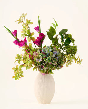 A white textured vase holds a bouquet of green hydrangeas, leafy branches and bright magenta silk flowers against a plain light background, creating a striking 'Cottage Garden' bouquet of artificial silk flowers that will add lasting charm to any space.