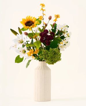 In a cream-colored vase is the Silk artificial flower bouquet 'Mom's Garden', an arrangement of sunflowers, lilies, white and burgundy flowers and green leaves against a plain background - making it look like an artificial flower bouquet reminiscent of a summer garden border.