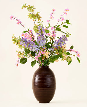 In a dark brown vase is a bouquet of pink, purple and yellow artificial silk flowers, the 'Sky Kiss' bouquet, with green leaves, against a plain light background. This maintenance-free decoration provides lasting beauty in any room, without the need for maintenance.