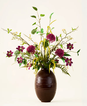 In a brown, ribbed vase is the bouquet "Woodland Charm" of silk artificial flowers, with purple alliums, dark pink flowers, yellow twigs and green leaves against a plain, light background.