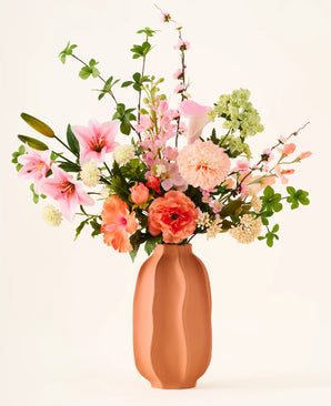 In a terracotta vase is the colorful bouquet 'Yasmine's Choice' of silk artificial flowers, consisting of pink lilies, peach roses and green leaves, made from high-quality silk flowers from Easyplants, all beautifully arranged against a light background.