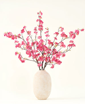 The Silk artificial flower bouquet Cherry blossom fuchsia 150 cm features vibrant fuchsia cherry blossom stems with green leaves in a simple white vase, which will complement any interior against a light background.