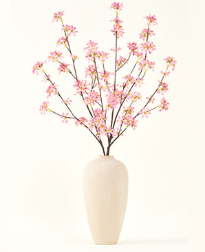 A tall beige ceramic vase contains several branches from the Silk artificial flower bouquet Pink flowers, with delicate pink blossoms and green leaves, against a plain white background.