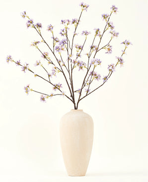 A tall, cream-colored ceramic vase contains a number of thin branches with the text Silk artificial flower bouquet Artificial flower branch purple, against a plain, light background.