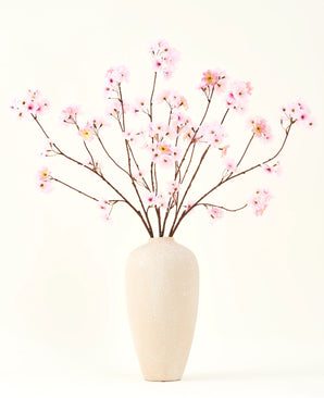 The Silk artificial flower bouquet Cherry blossom pink 114 cm has long, thin branches with bunches of light pink flowers in a beige ceramic vase, which form a striking contrast against the plain white background.