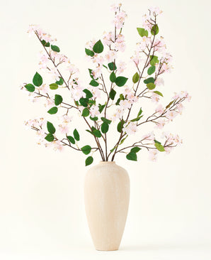 A beige ceramic vase contains several long branches with the Silk artificial flower bouquet of Peach Blossom pink and green leaves, against a plain white background - perfect for an artificial floral interior.
