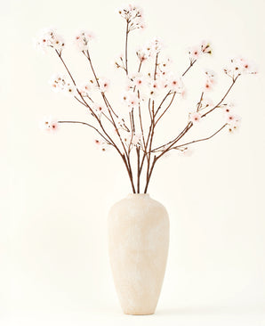 The Silk artificial flower bouquet Cherry blossom blush 114 cm has tall, delicate branches in a simple beige vase, which adds a soft, elegant touch to your interior against a plain, light background.