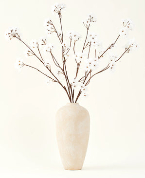 The Silk artificial flower bouquet Cherry blossom cream 114 cm stands tall in a beige ceramic vase and brings a serene decoration to any room with its delicate, cream-colored branches against a light background.