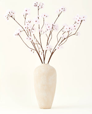 The Silk artificial flower bouquet Cherry blossom light pink 114 cm stands in a tall beige ceramic vase with long, thin branches and delicate light pink flowers, creating a serene artificial floral accent against a plain background.