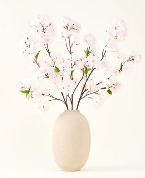 The Silk artificial flower bouquet Cherry blossom blush 119 cm shows delicate, light pink cherry blossoms with green leaves in a beige vase, ideal as an artificial flower decoration against a light background.