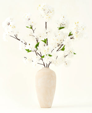 The Silk artificial flower bouquet Cherry blossom cream 119 cm has elegant brown branches with soft white fake blossoms and green leaves, displayed in a simple beige vase against a plain light background.