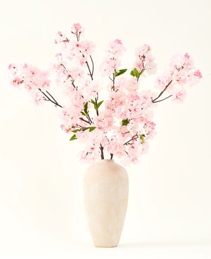 The Silk artificial flower bouquet Cherry blossom pink 119 cm, arranged in a beige ceramic vase with delicate green leaves, creates a serene interior accent against a simple light background.