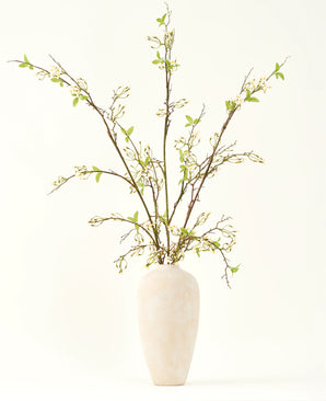 A tall, cream-colored ceramic vase contains the Silk artificial flower bouquet Verbena cream with long branches, small green leaves and delicate white blossoms, beautifully arranged against a plain light background.