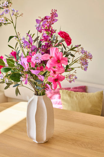 In a white ceramic vase is the 'Purple Dawn' bouquet of silk artificial flowers, a lively arrangement of pink, purple and magenta flowers; the vase stands on a light wooden table with colorful pillows in the background.