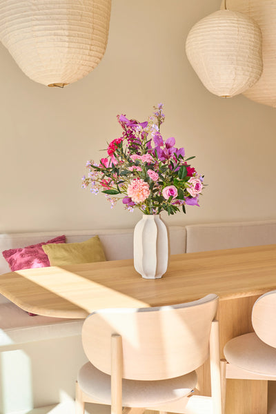 On a light wooden dining table sits a white vase with the Purple Dawn silk artificial flower bouquet, bursting with vibrant pink and purple hues. Behind it, an upholstered bench with cushions and two hanging paper lanterns complete the scene, imbuing the space with the tranquil atmosphere inspired by Purple Dawn.