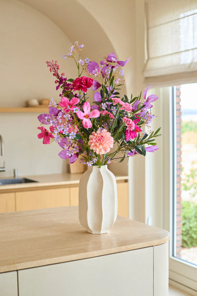 On a kitchen island made of light wood there is a white vase with the 'Purple Dawn' bouquet of artificial silk flowers - consisting of pink and purple artificial flowers - with a window and beige kitchen cabinets in the background.