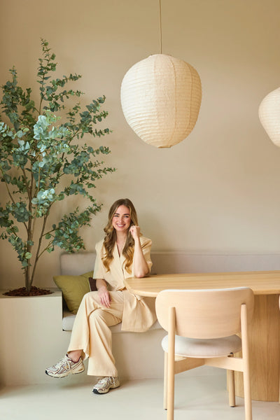 A woman in a beige suit sits smiling at a wooden table in a modern, minimalist room with light walls, paper pendant lamps hanging from the ceiling and a 180 cm Artificial Eucalyptus plant that adds some greenery to the room.